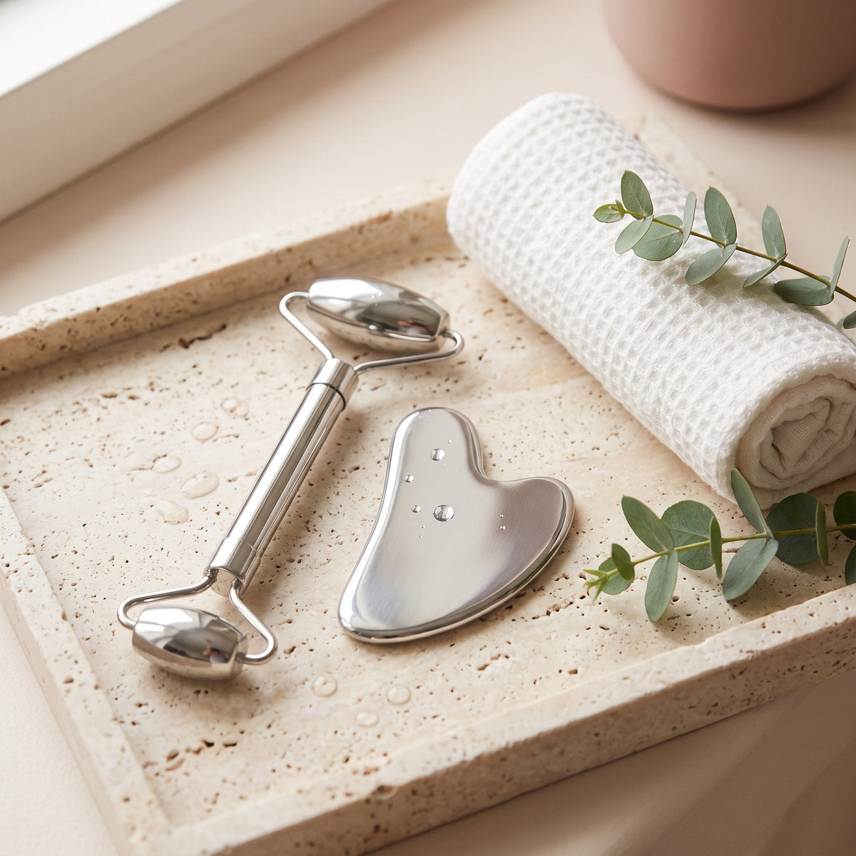 Stainless steel face roller and heart-shaped gua sha on a travertine tray with a rolled towel